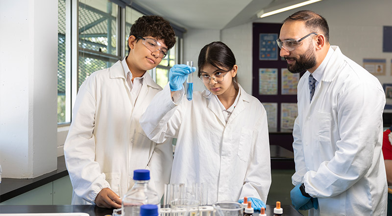 Science teacher at Delany College Granville assisting students with lab experiment. All are wearing coats and goggles